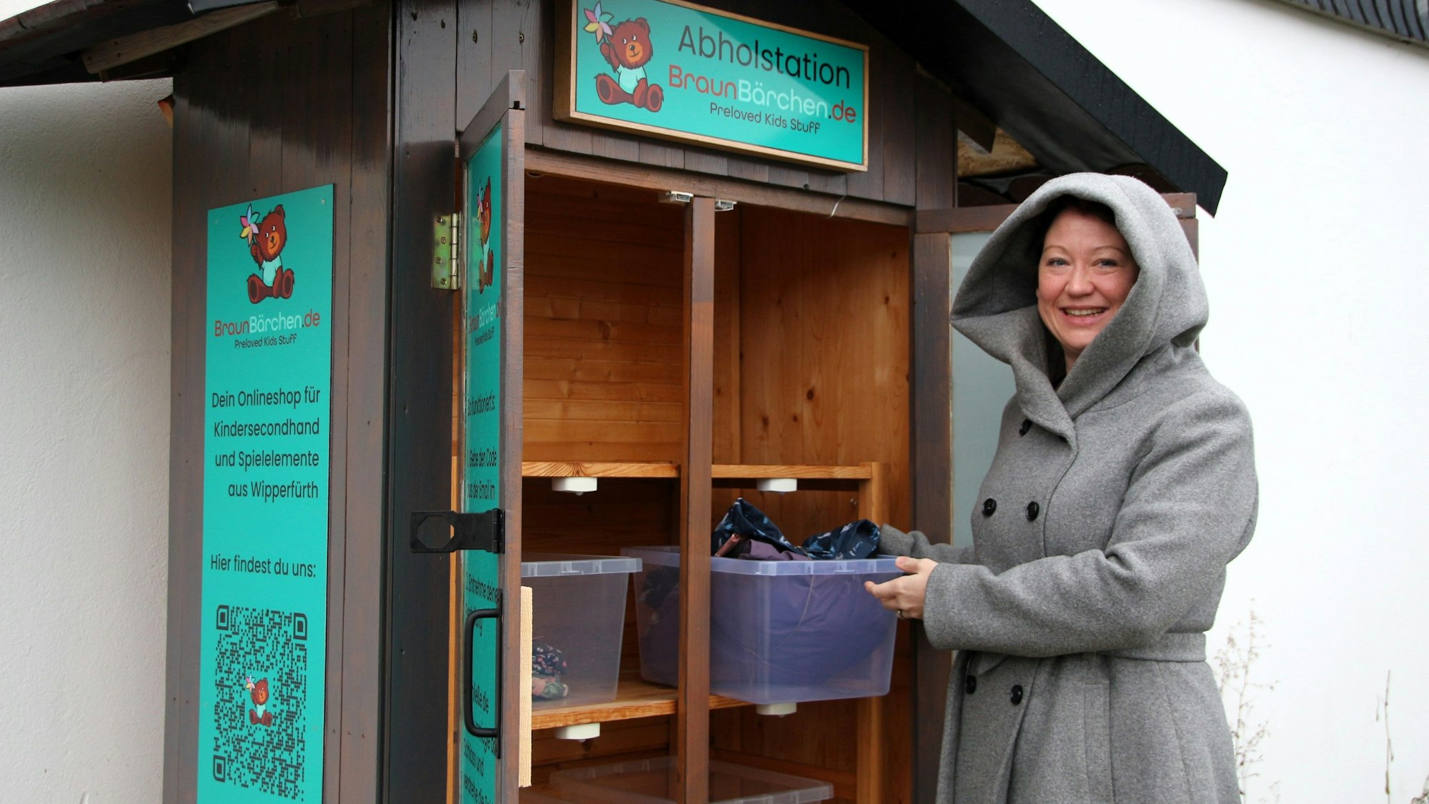 Sina Braun steht vor einem Außenschrank, in dem große Plastikboxen stehen. Sie trägt einen Mantel mit Kapuze und schaut in die Kamera.
