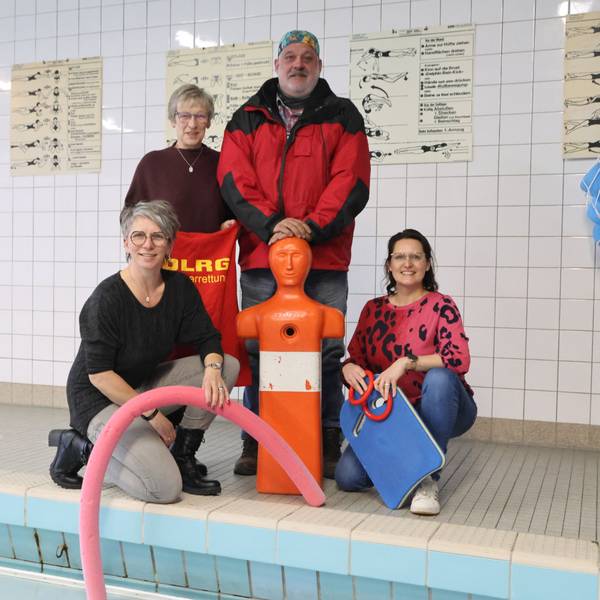 Eine Gruppe Männer und Frauen, unter anderem von der DLRG, stehen mit einer Trainingspuppe am Beckenrand eines Hallenbads.