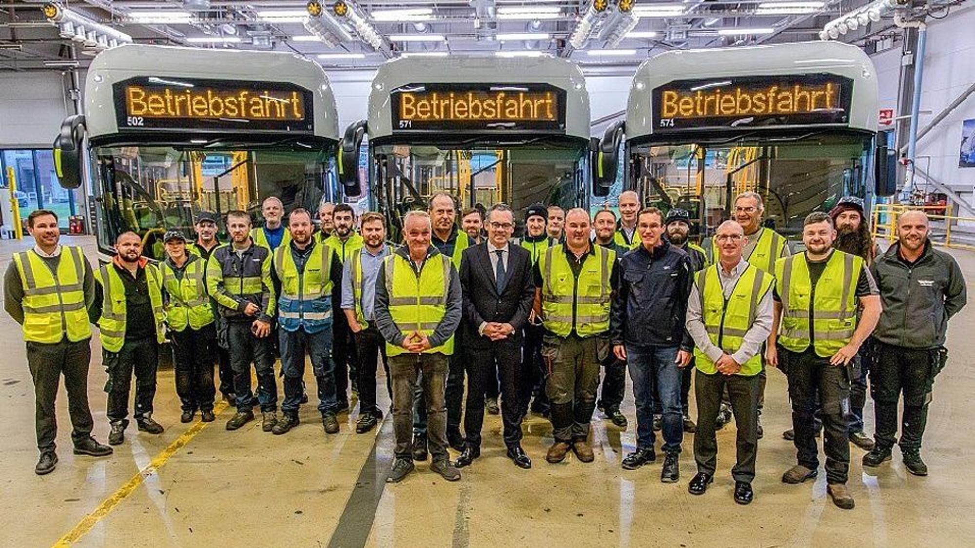 Auf dem Foto sind Männer in Anzügen und andere in gelben Warnwesten vor Bussen zu sehen.