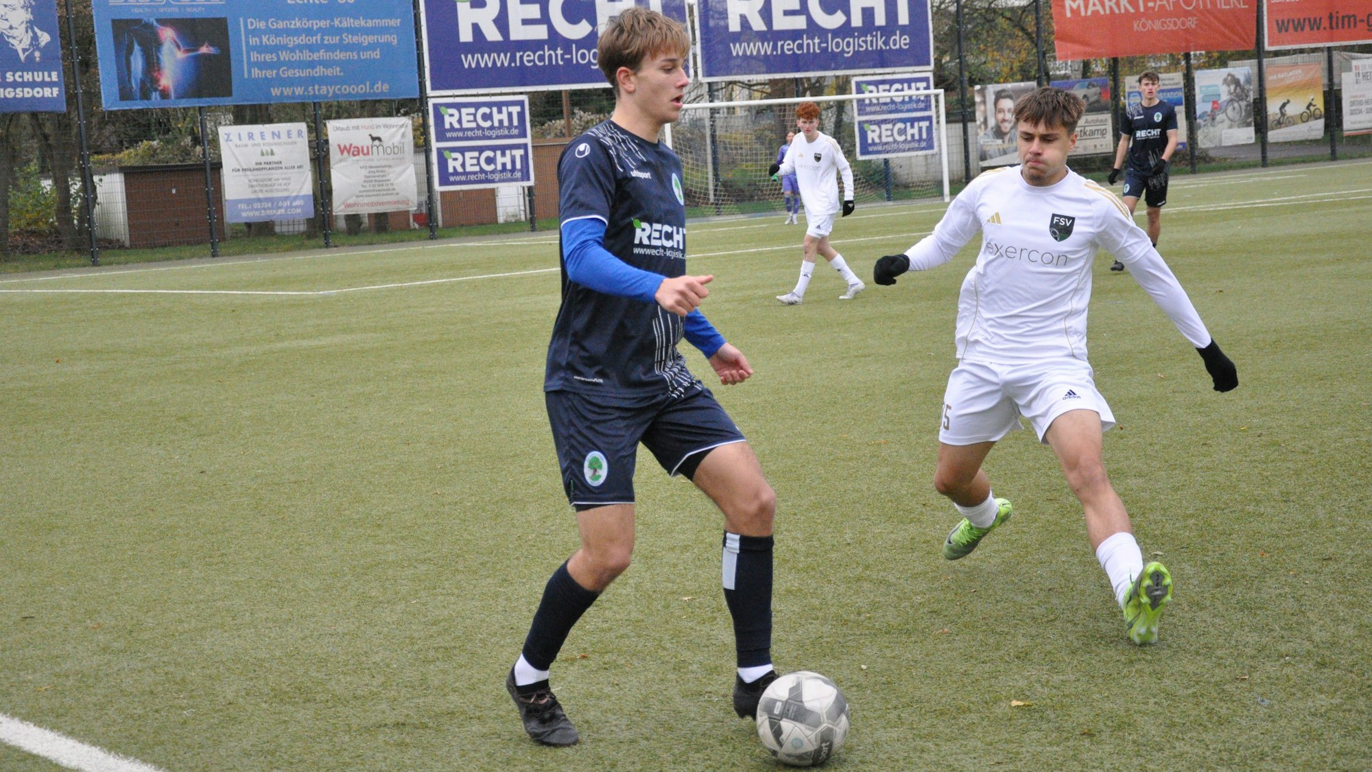 Das Bild zeigt einen Spieler im blauen Trikot, der den Ball vor seinem Gegner im weißen Dress dem Ball behauptet.