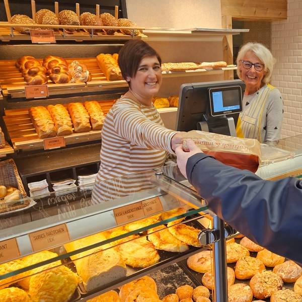 Zu sehen sind zwei Frauen hinter der Verkaufstheke einer Bäckerei, eine der beiden reicht einer Kundin eine Tüte mit Brötchen.