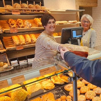 Zu sehen sind zwei Frauen hinter der Verkaufstheke einer Bäckerei, eine der beiden reicht einer Kundin eine Tüte mit Brötchen.