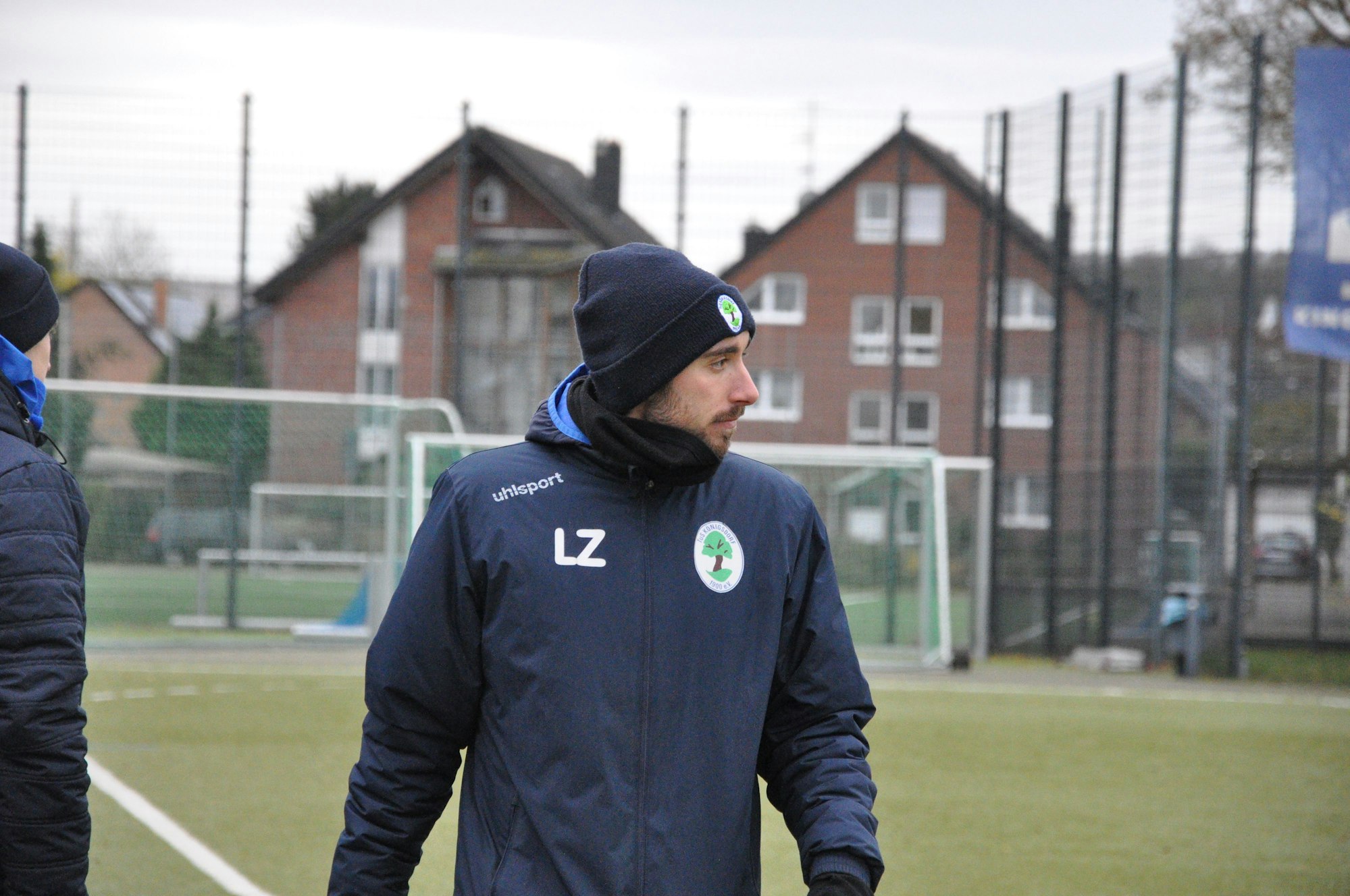 Das Bild zeigt einen in Blau gekleideten Trainer mit Mütze und Schal, der auf den Platz schaut.