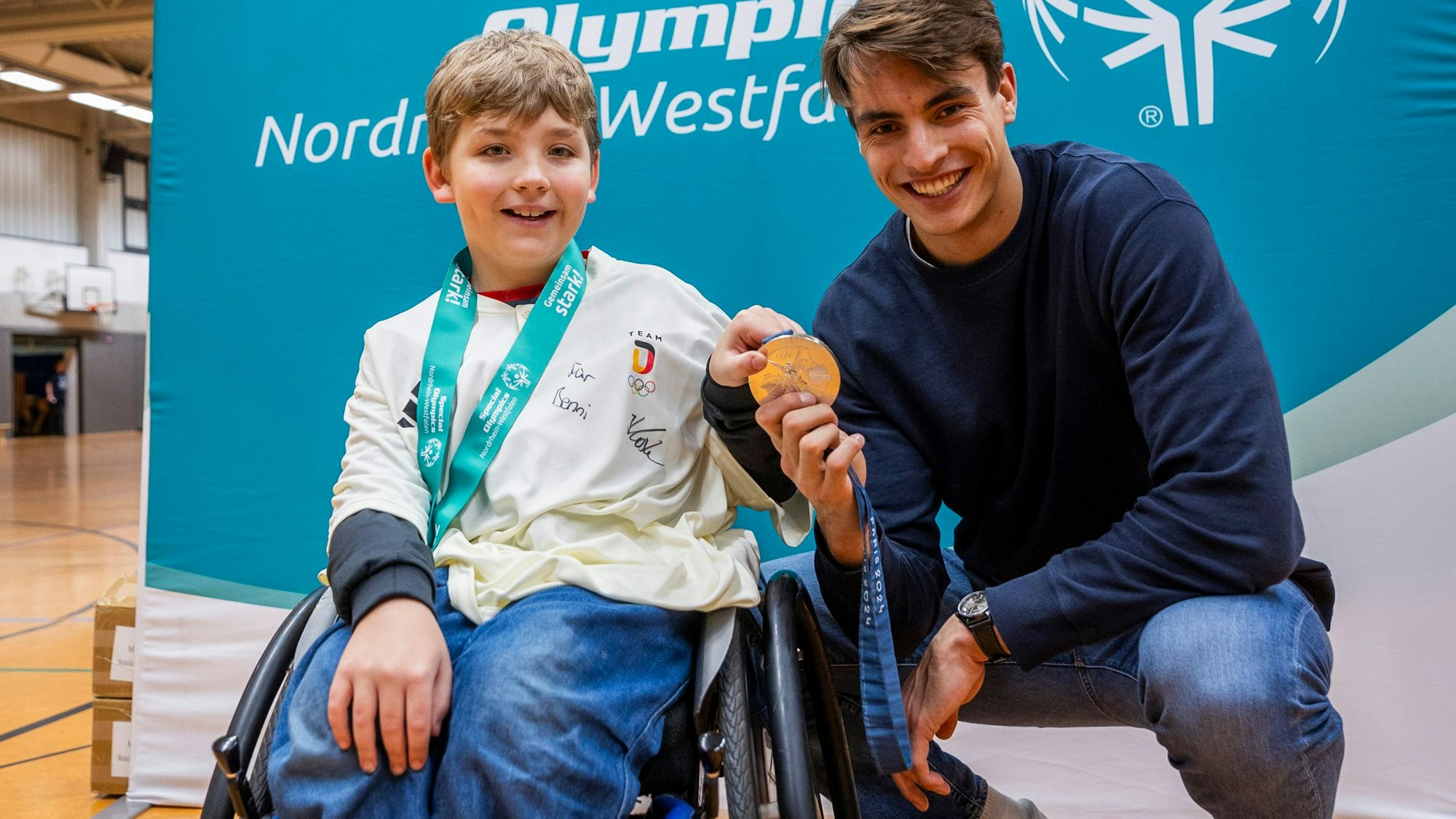 Beim Special-Olympics-Handball-Turnier in Hamm im Dezember trafen sich Julian Köster (r.) und Benni (l.) schließlich wieder. Sie halten gemeinsam eine olympische Medaille in Richtung der Kamera, lächeln. Benni sitzt im Rollstuhl.