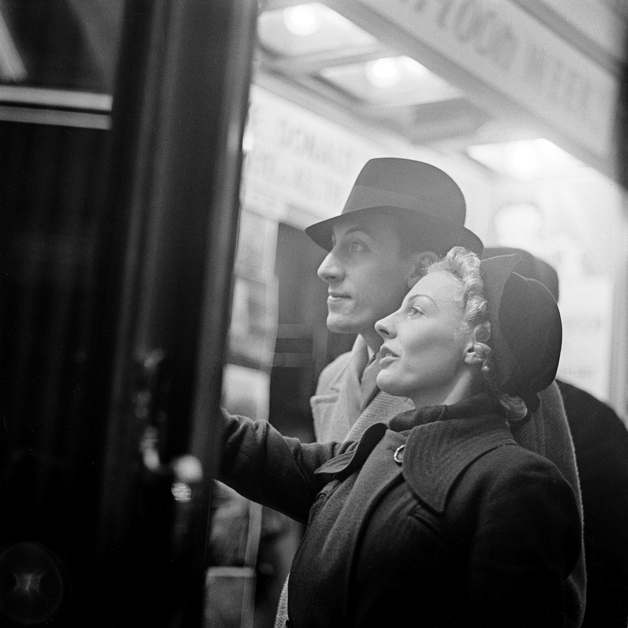 Auf einer älteren s/w-Aufnahme sind ein Mann und eine Frau vor einem erleuchteten Schaufenster zu sehen. Beide tragen Mantel und Hut.