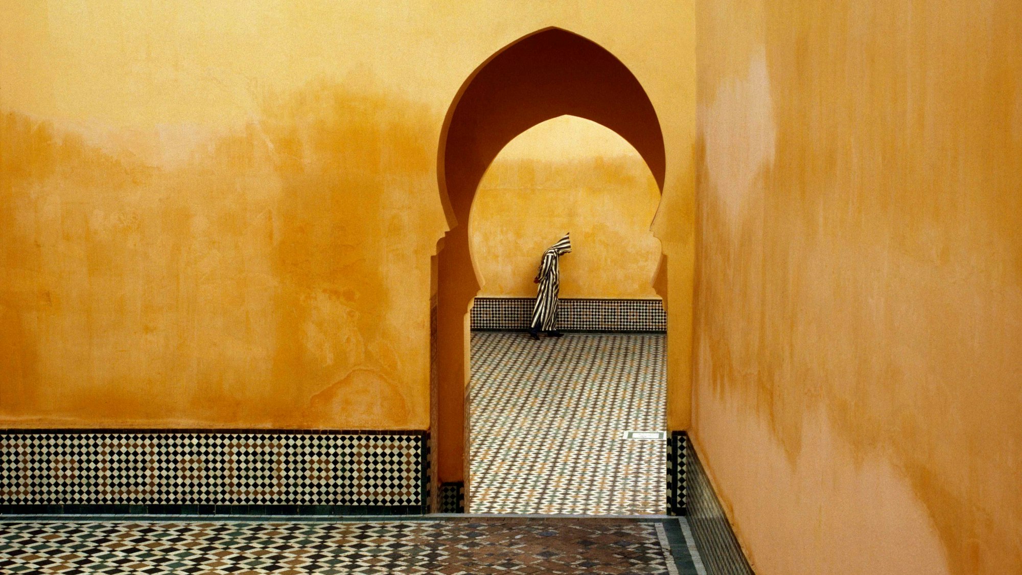 Die Aufnahme „MOROCCO. Meknes. Moulay Ismael Mausoleum (Muslim shrine). 1985“ zeigt den Blick durch ein Tor, im Hintergrund steht ein Mann mit einer gestreiften Kutte mit Kapuze.