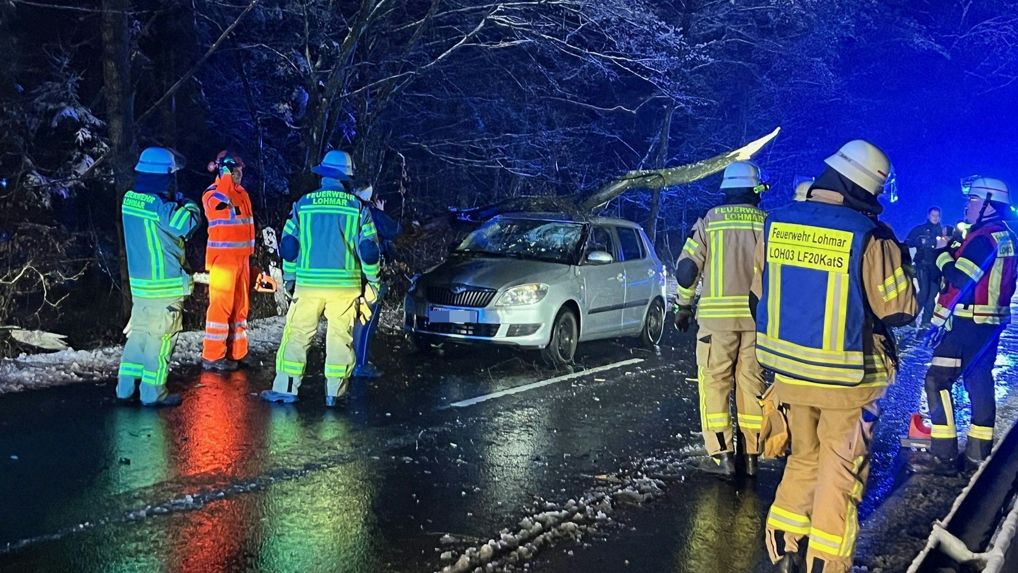Feuerwehrleute auf einer verschneiten Straße. Auf einem Pkw liegt ein großer Baum.