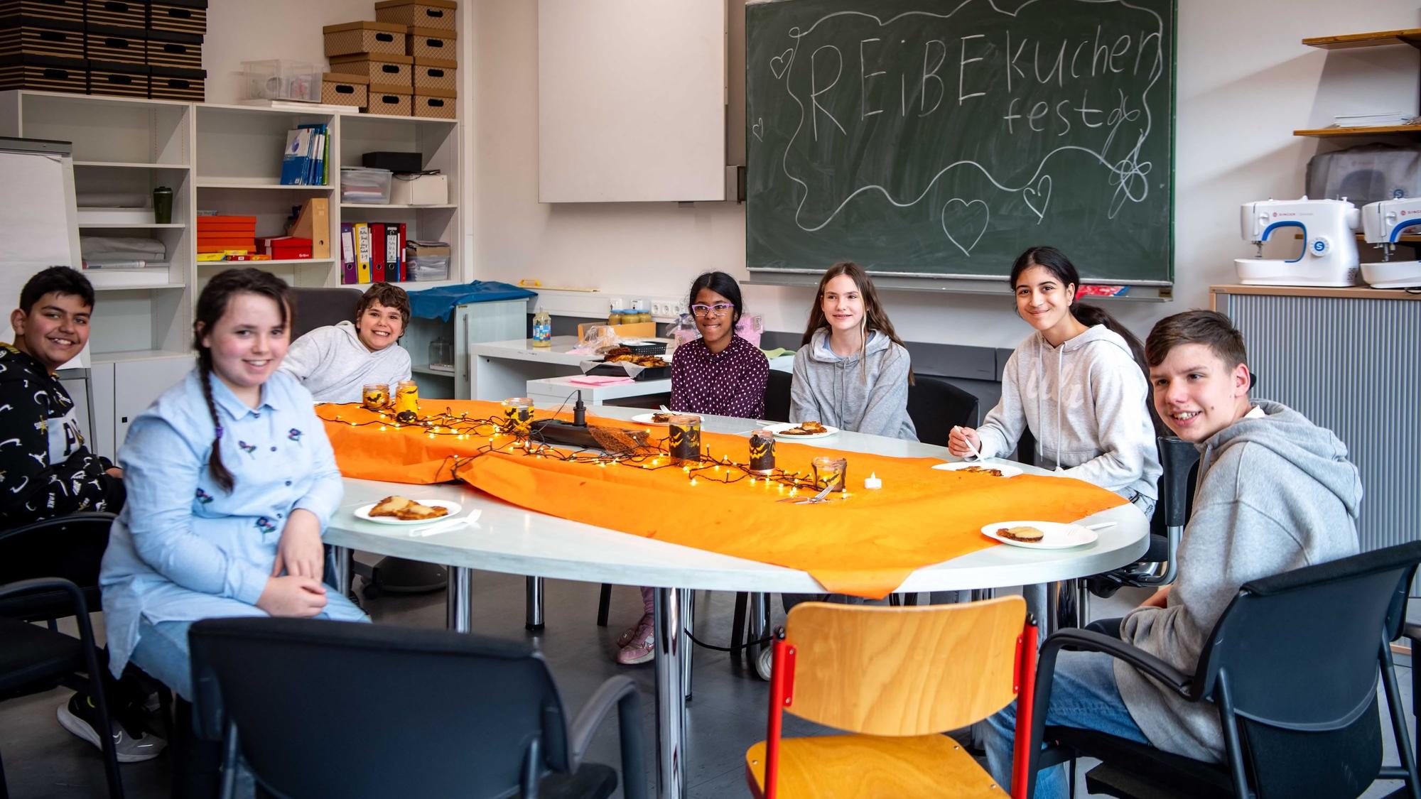 Mehrere Schüler und eine Lehrerin sitzen an einem ovalen Tisch und haben auf Tellern Reinekuchen vor sich liegen.