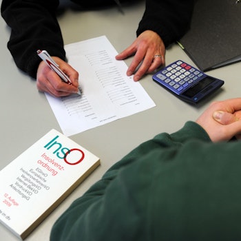 Das Symbolbild zeigt eine Beratungssituation. Vor dem Berater liegen ein Buch, eine Liste und ein Taschenrechner.