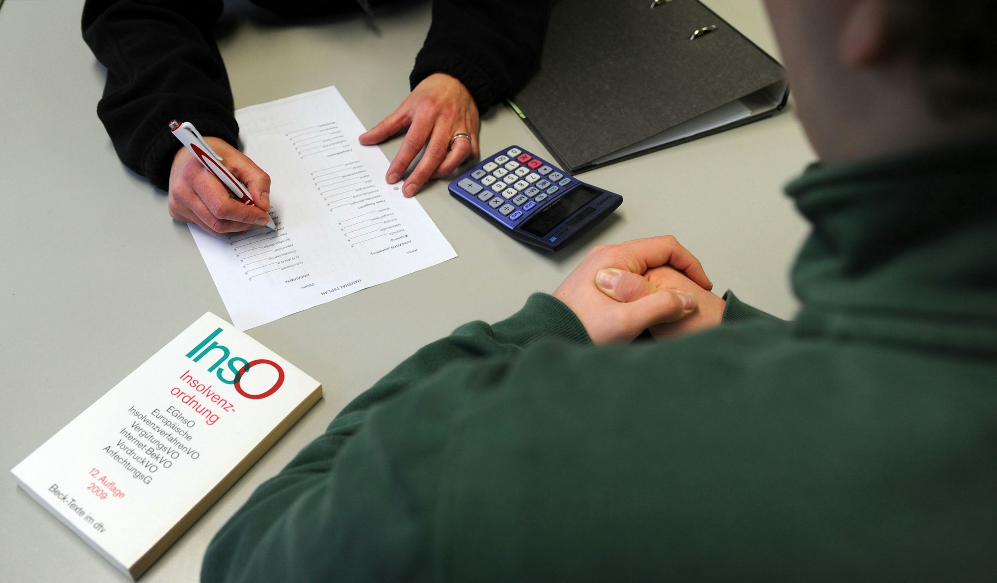 Das Symbolbild zeigt eine Beratungssituation. Vor dem Berater liegen ein Buch, eine Liste und ein Taschenrechner.