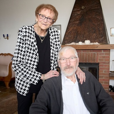 Marliese Hück steht hinter ihrem Mann Klaus, der im Wohnzimmer sitzt.