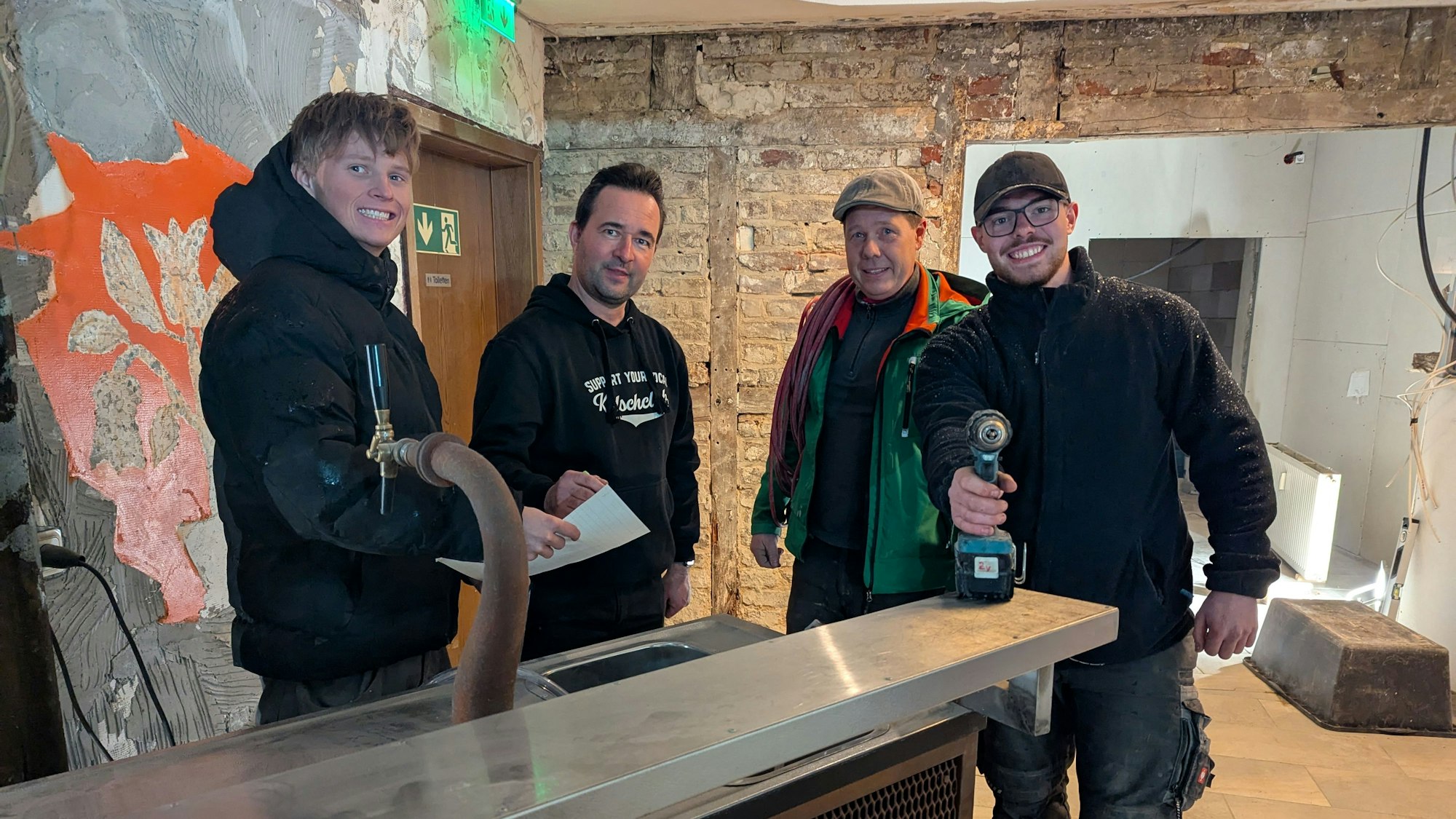 Vier Männer stehen, teils mit Werkzeug in der Hand, hinter einem Tresen vor kahlen Wänden.