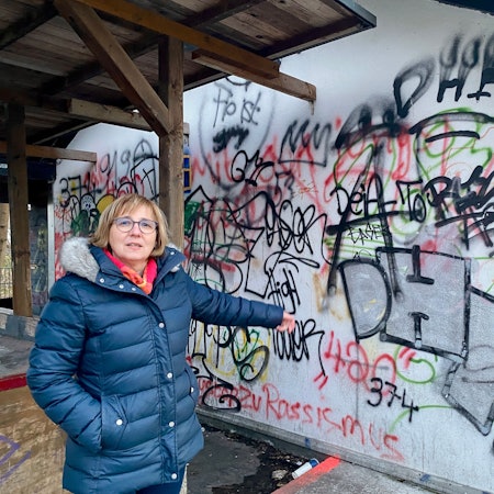 Eine Frau in einer dunkelblauen Steppjacke zeigt auf eine beschmierte Wand.