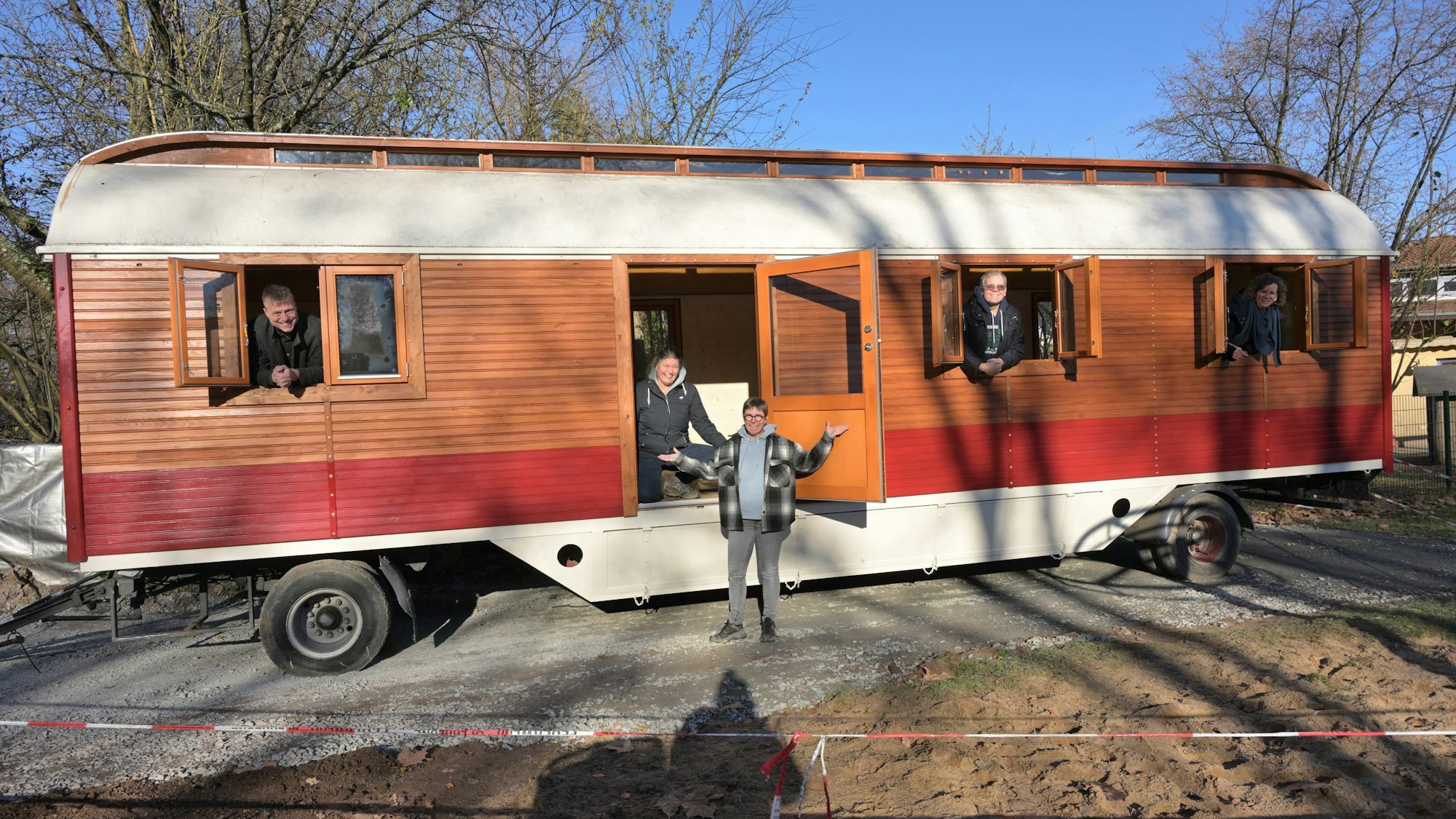 Ein umgebauter Zirkuswagen steht auf einem Schotterplatz. Fünf Personen schauen aus den Fenstern und der Tür.