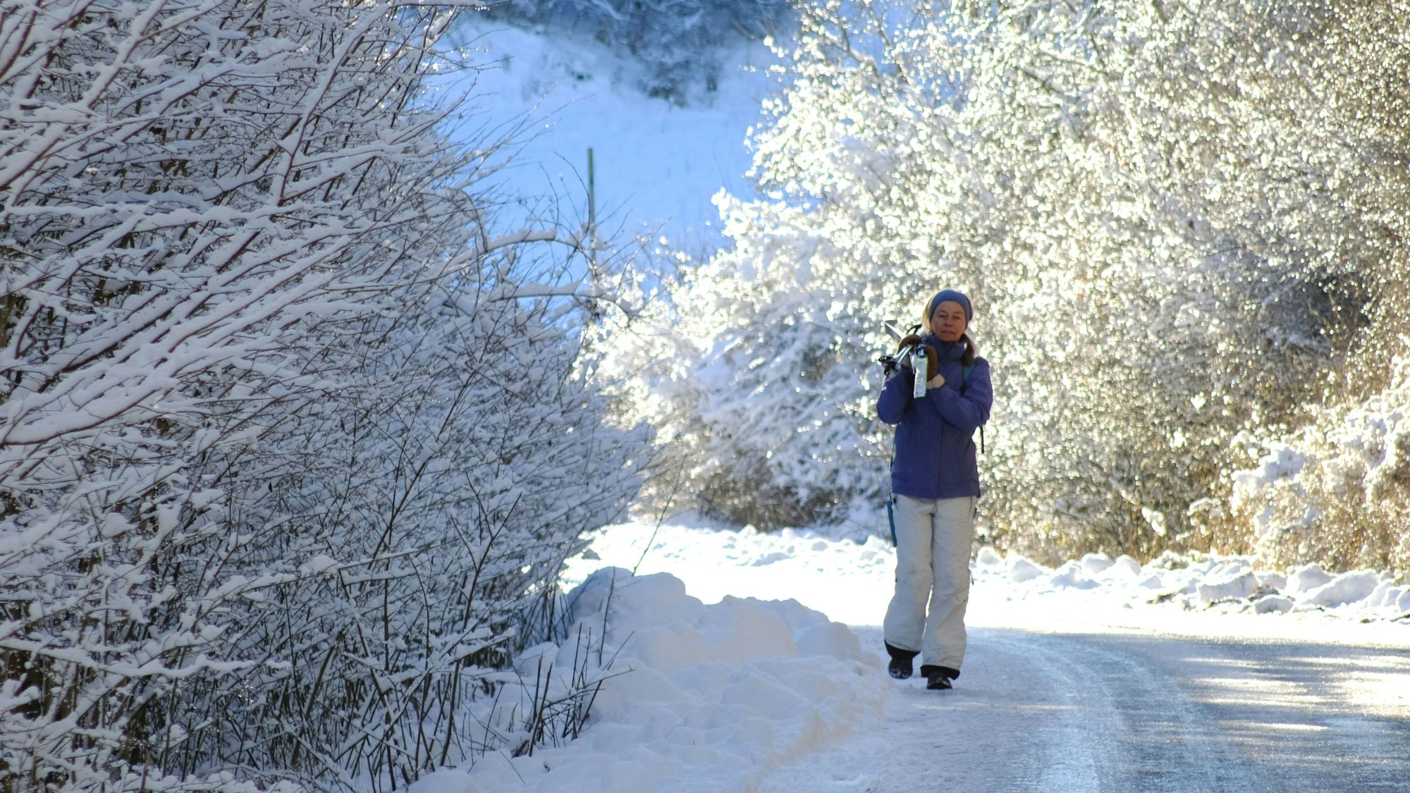 Eine Spaziergängerin im verschneiten Urfttal hat ihre Langlaufskier geschultert.
