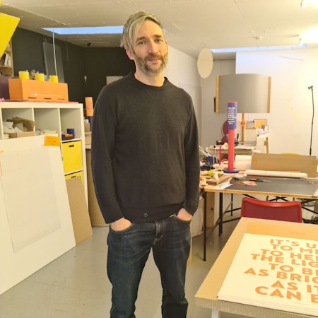 Ein Mann mit grauem Haar und Bart steht in einer Grafiker-Werkstatt neben einem Tisch.