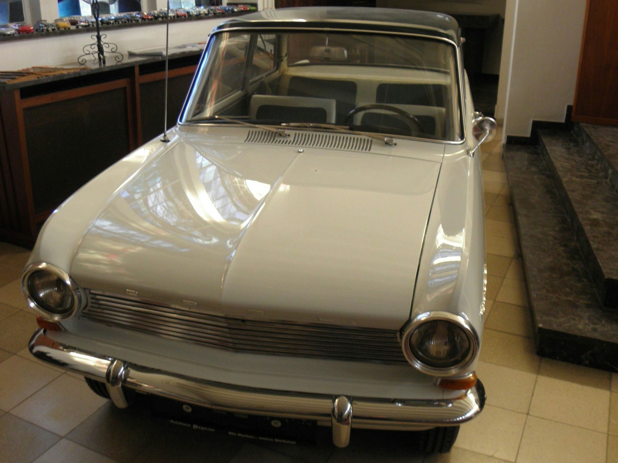 Ein altes weißes Auto, ein Opel Kadett, steht in einem Raum mit einem gefliesten Boden.