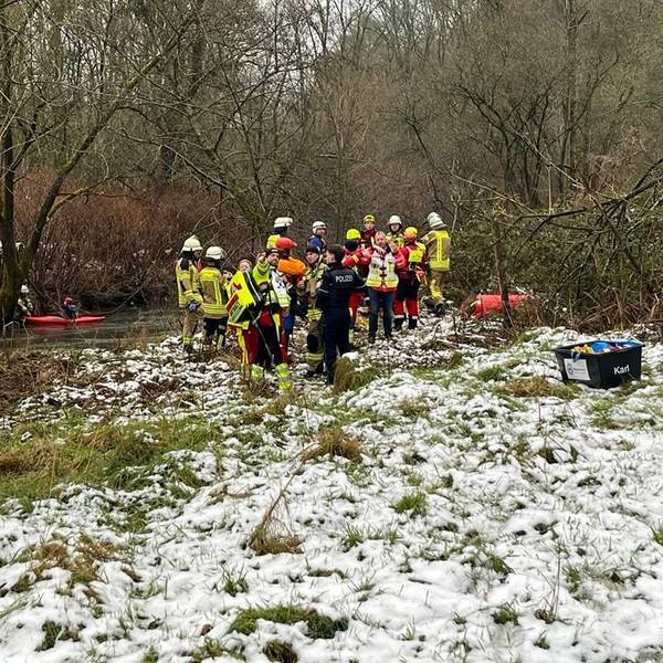Rettungskräfte stehen an der Sülz auf einer schneebedeckten Wiese.