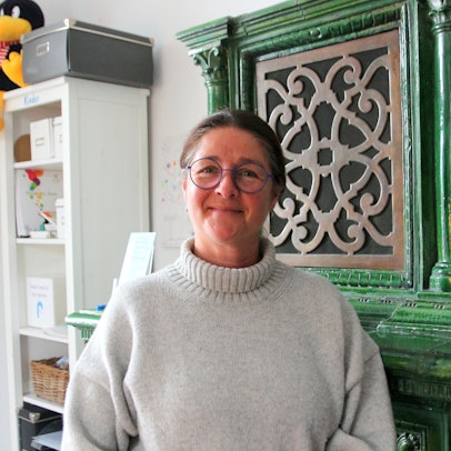 Auf dem Foto ist eine Frau zu sehen. Sie trägt einen hellen Rollkragenpullover und Brille. Sie steht vor einem antiken grünen Schrank.