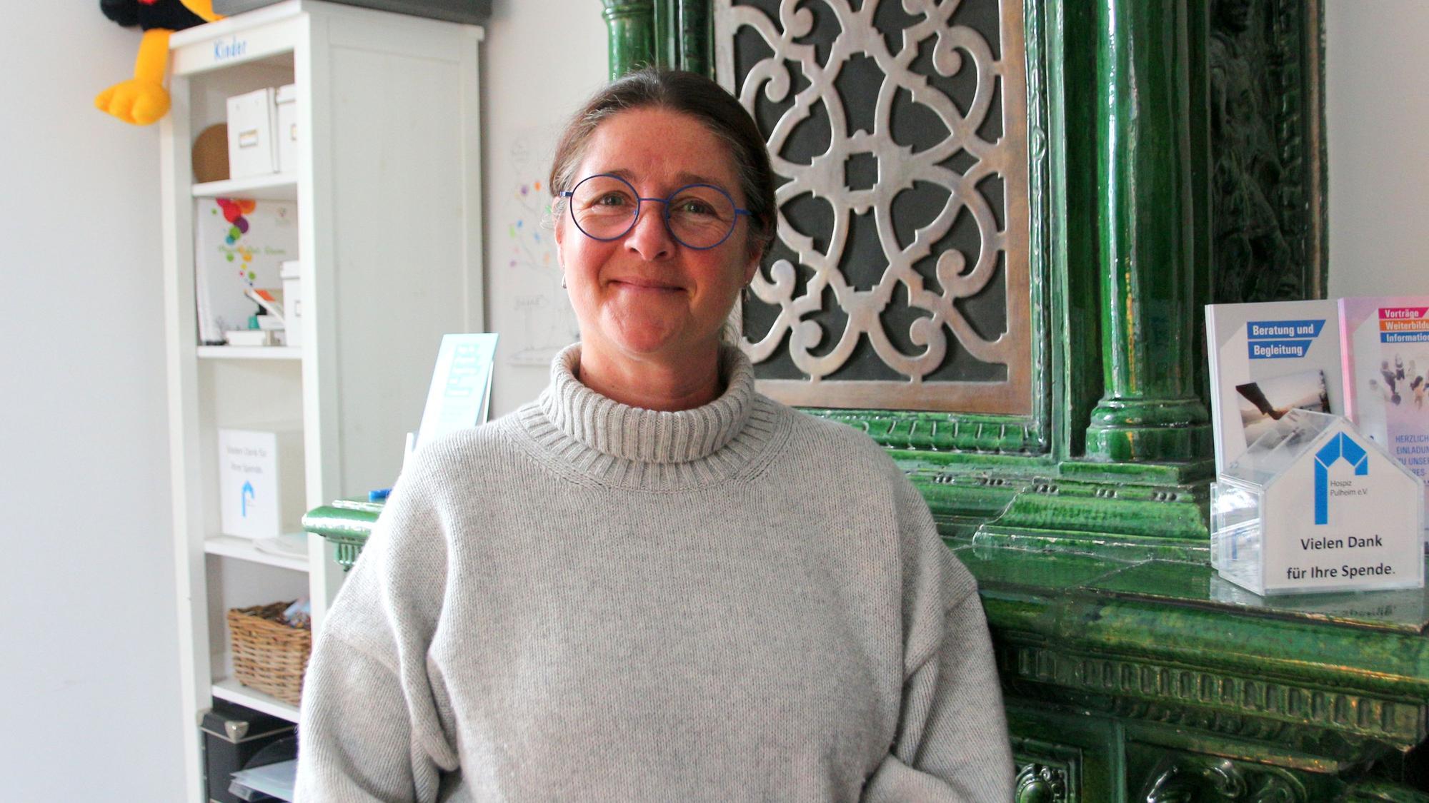 Auf dem Foto ist eine Frau zu sehen. Sie trägt einen hellen Rollkragenpullover und Brille. Sie steht vor einem antiken grünen Schrank.