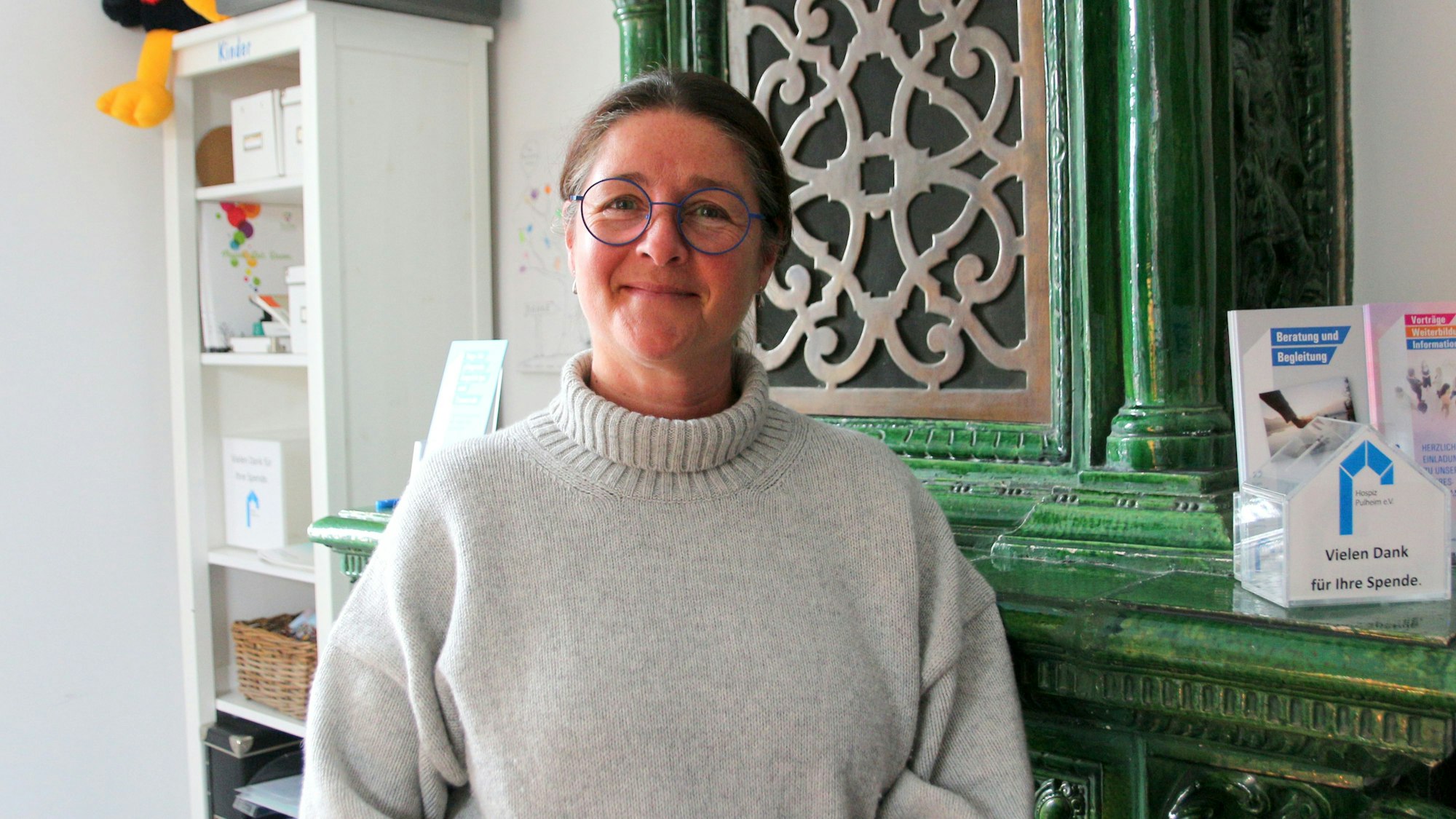 Auf dem Foto ist eine Frau zu sehen. Sie trägt einen hellen Rollkragenpullover und Brille. Sie steht vor einem antiken grünen Schrank.