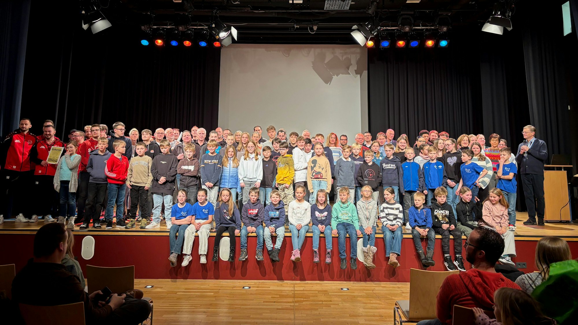 Zahlreiche Erwachsene und Kinder stehen auf einer Bühne für ein Gruppenfoto.