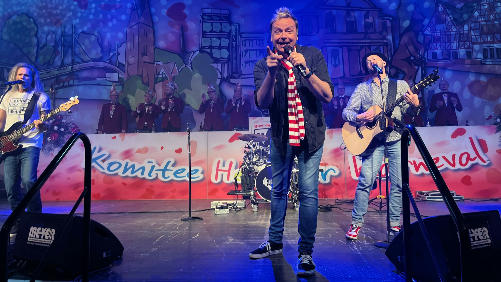 Drei Musiker stehen auf einer Bühne, der Sänger zeigt ins Publikum.