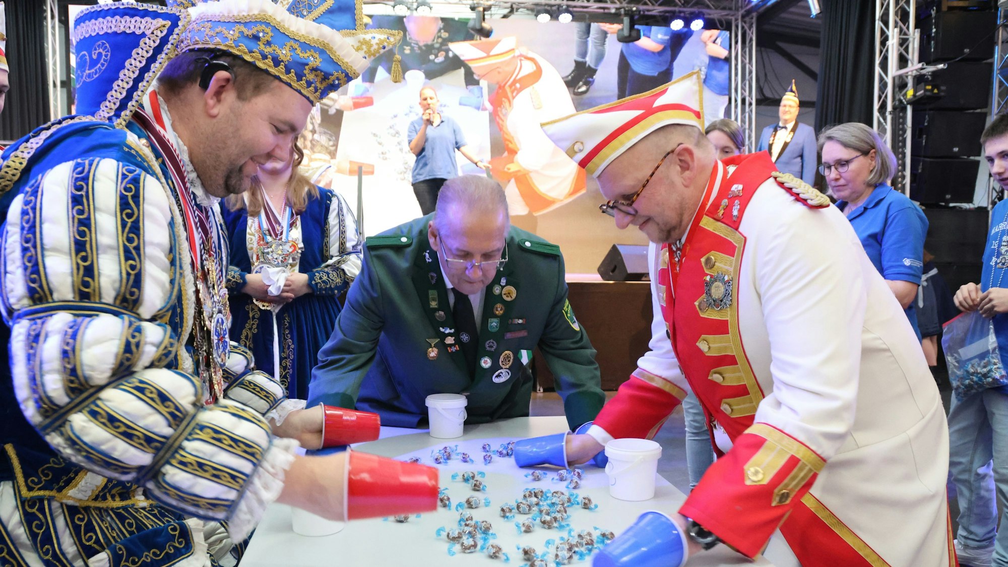 Ein Prinz einer Karnevalsgesellschaft sammelt mit Bechern um seine Hände Bonbons auf einem Tisch ein.