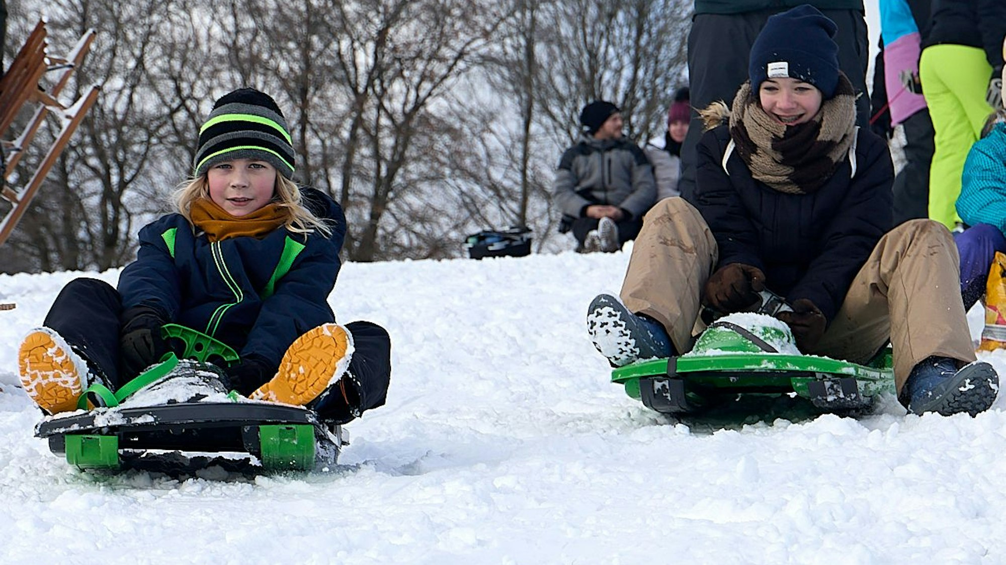 Zwei Kinder sitzen auf Rodelschlitten und fahren einen verschneiten Hang hinab.