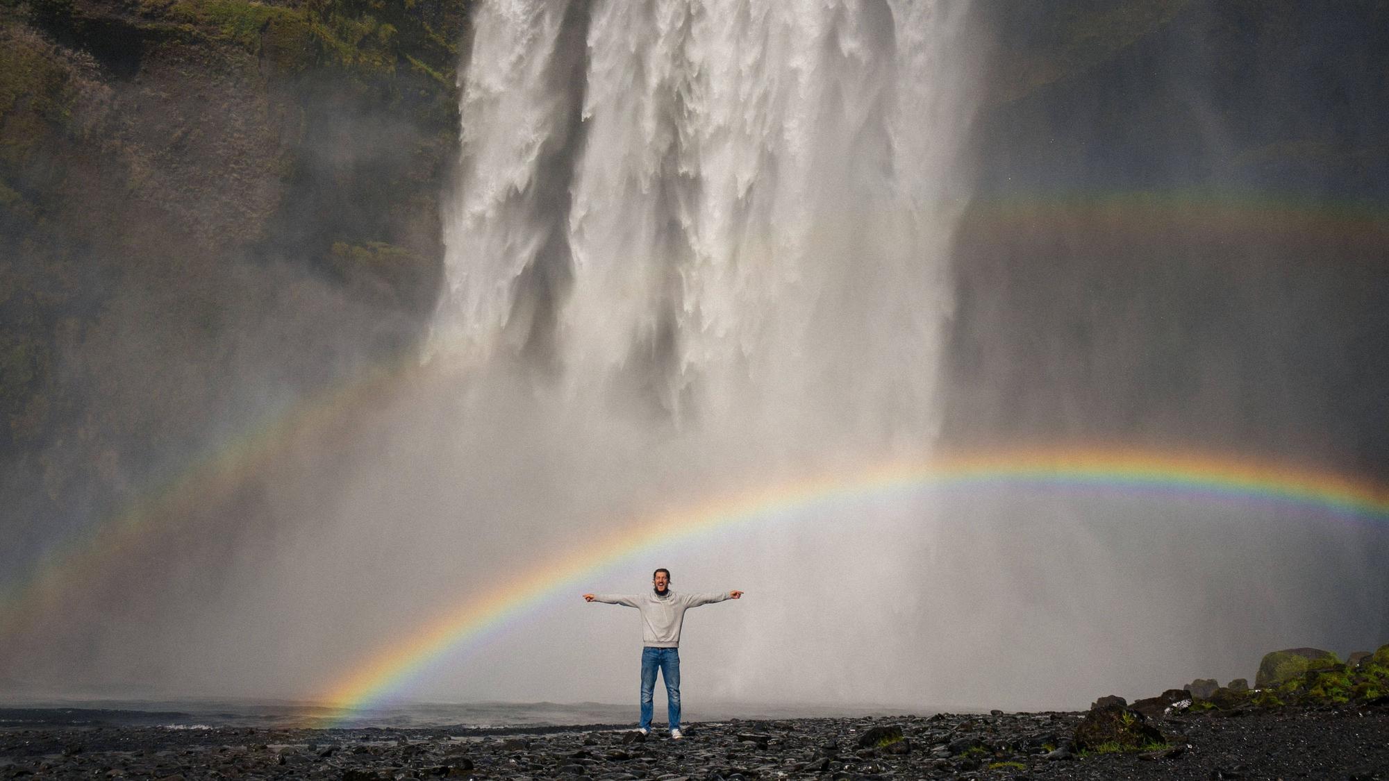 Ein Mann steht mit ausgestreckten Armen vor dem Wasserfall Skogafoss.