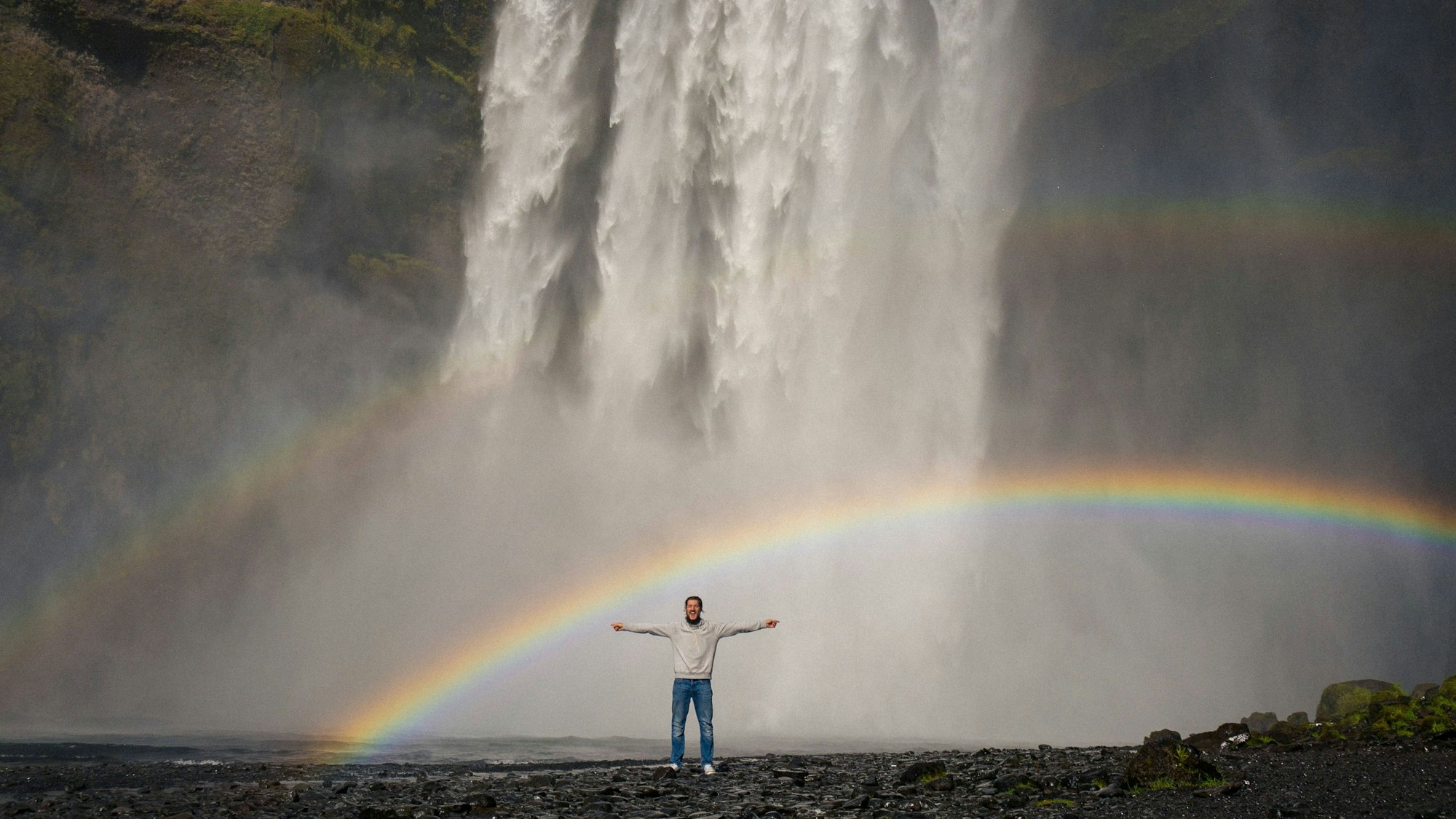 Ein Mann steht mit ausgestreckten Armen vor dem Wasserfall Skogafoss.