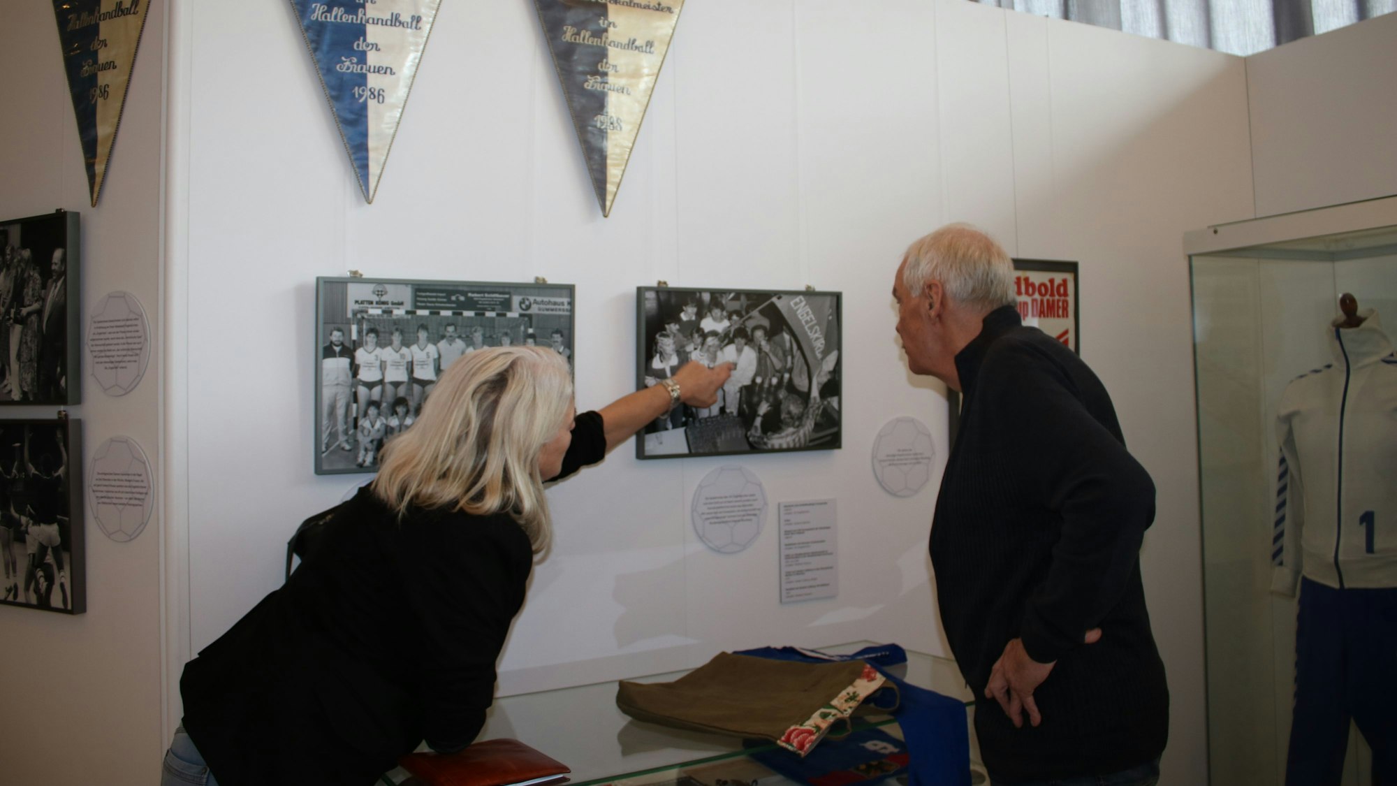Eine Frau zeigt in einer Ausstellung auf ein altes Foto an der Wand. Ein Mann steht neben ihr und schaut ebenfalls darauf.