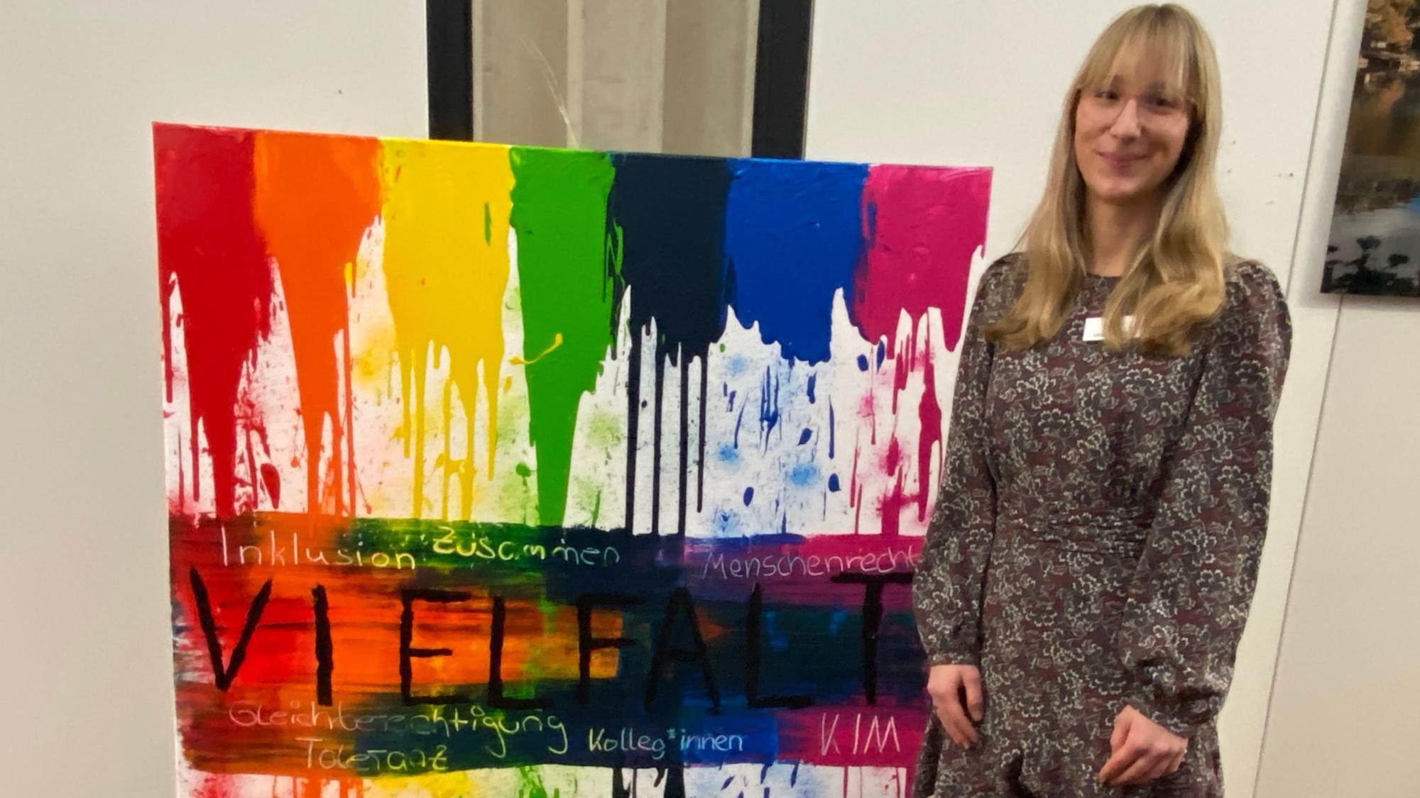 Eine junge Frau steht rechts von einem bunten Gemälde, das zum Thema Vielfalt gemalt wurde.