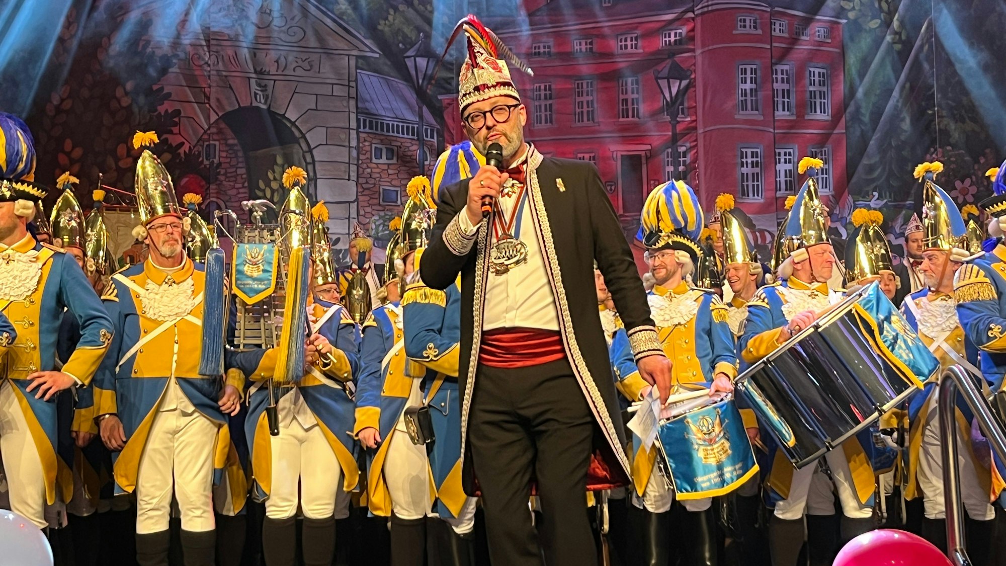 Der Sitzungspräsident einer Karnevalsveranstaltung steht vor einem Traditionscorps auf der Bühne.