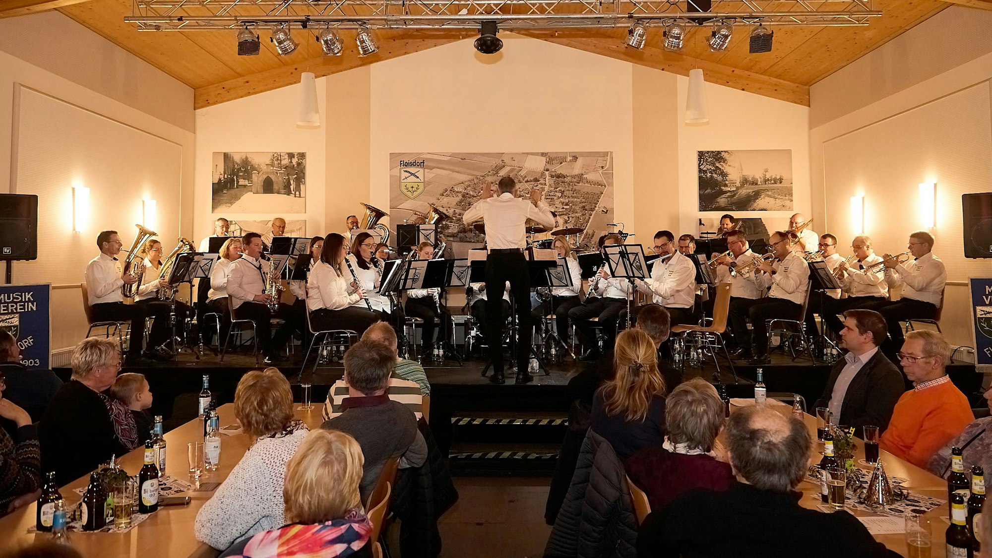 Im Dorfsaal Floisdorf stehen die Musiker des Musikvereins Cäcilia Floisdorf aufv der Bühne. Das Publikum sitzt an den Tischen und lauscht.
