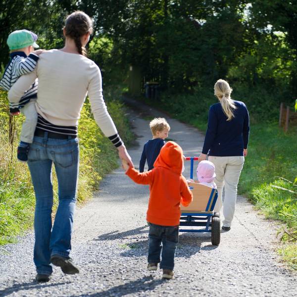 Eine Tagesmutter zieht einen Bollerwagen mit Kindern, dahinter läuft eine Tagesmutter mit einem Kind auf ihrem Arm.