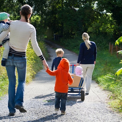 Eine Tagesmutter zieht einen Bollerwagen mit Kindern, dahinter läuft eine Tagesmutter mit einem Kind auf ihrem Arm.