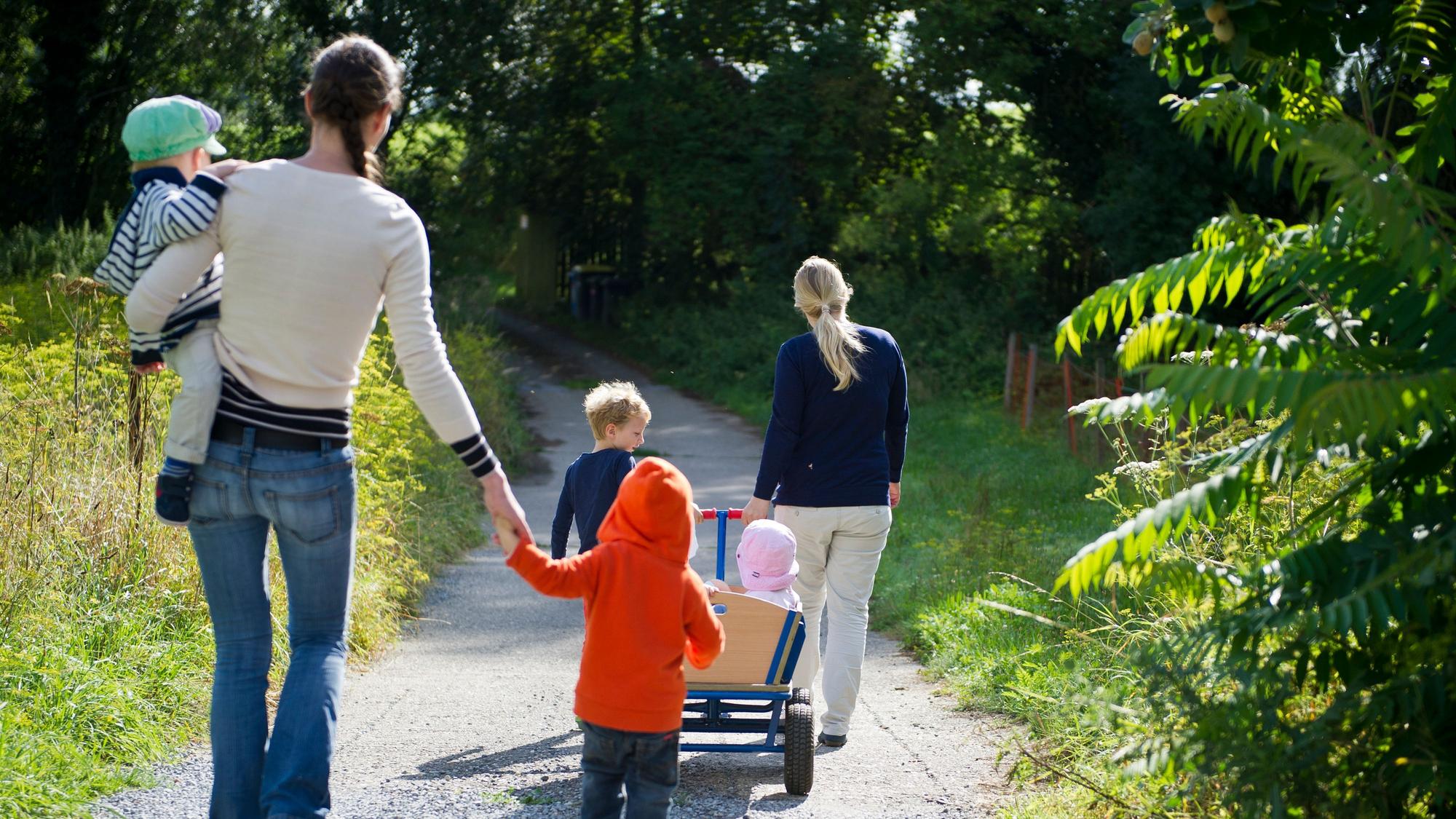 Eine Tagesmutter zieht einen Bollerwagen mit Kindern, dahinter läuft eine Tagesmutter mit einem Kind auf ihrem Arm.