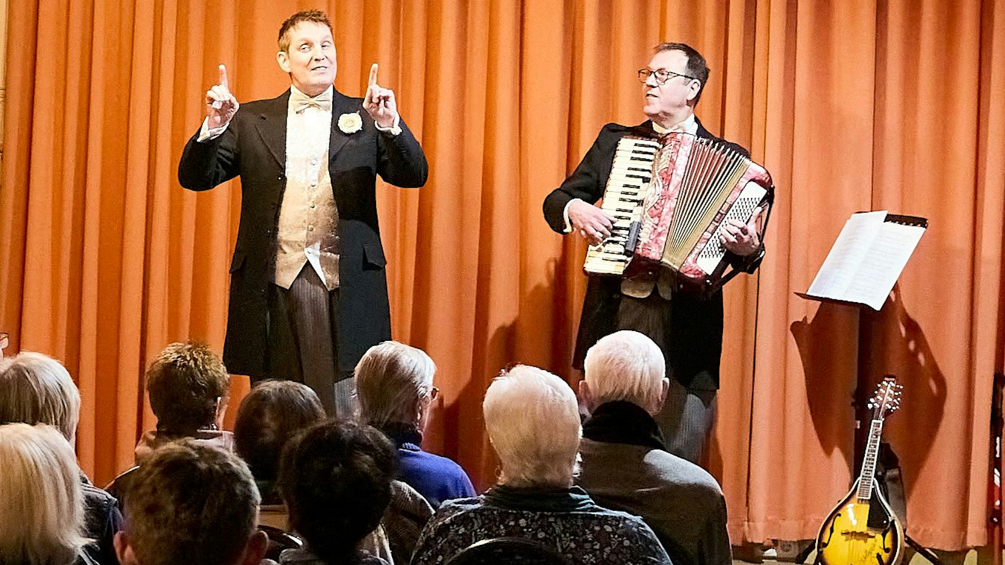 Zwei Männer im Frack, der eine mit Akkordeon, der andere mit zwei erhobenen Zeigefingern, stehen auf der Bühne.