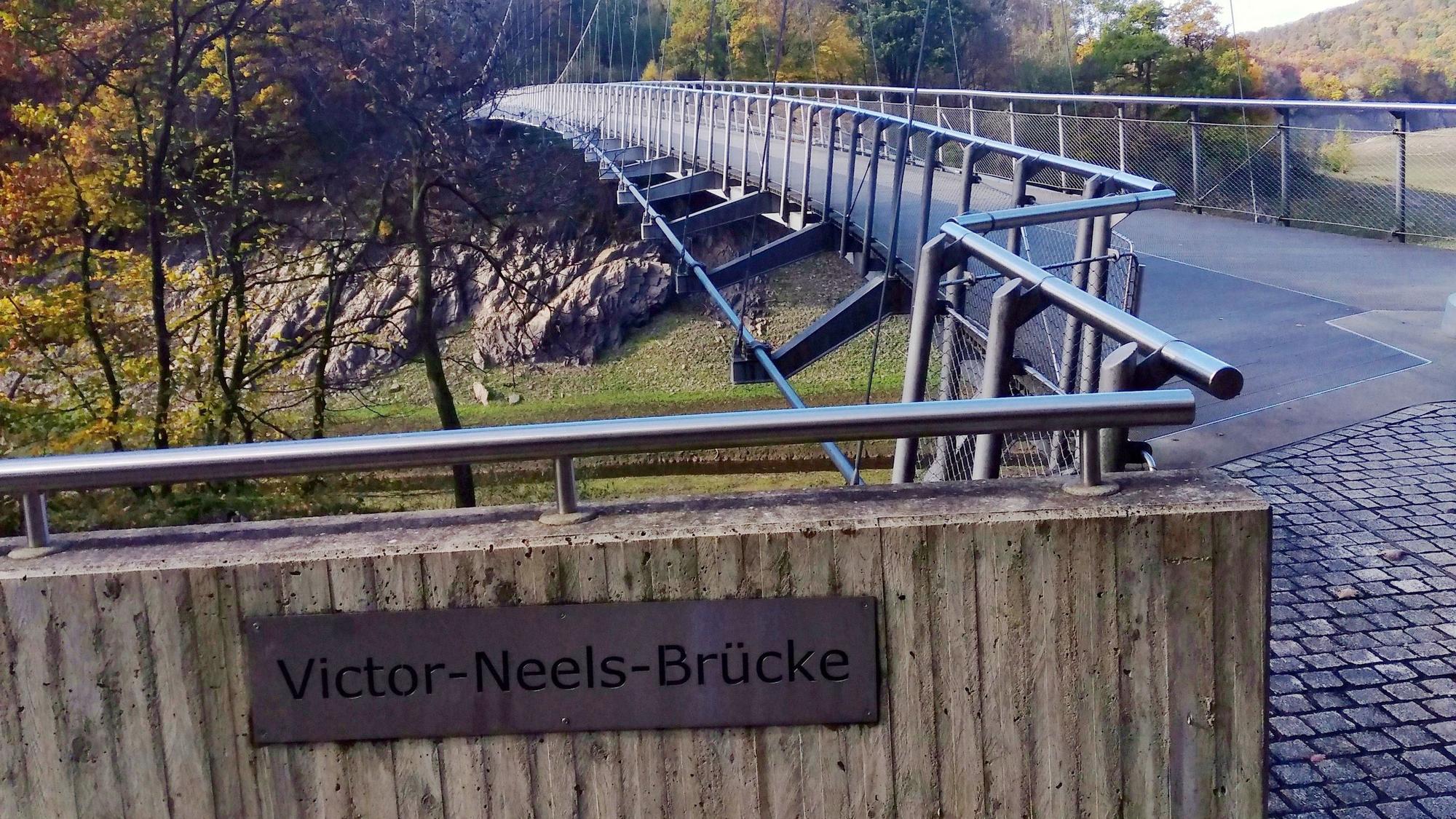 An der Brücke, die über den Urftsee führt, ist eine Plakette mit dem Schriftzug „Victor-Neels-Brücke“ angebracht.