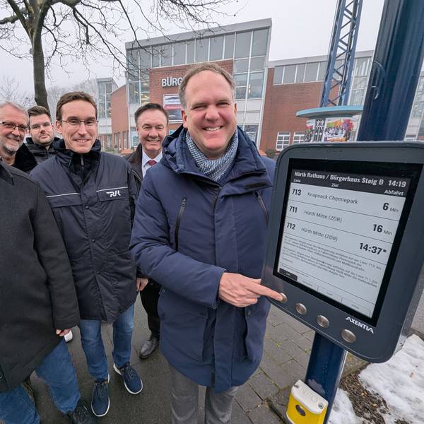 Das Foto zeigt eine Gruppe von Menschen vor dem digitalen Anzeigegerät an der Bushaltestelle.