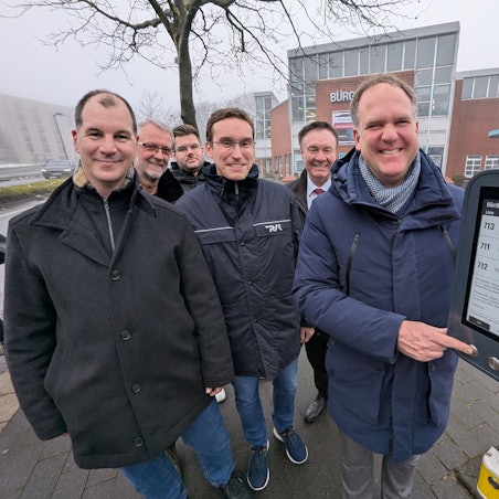 Das Foto zeigt eine Gruppe von Menschen vor dem digitalen Anzeigegerät an der Bushaltestelle.