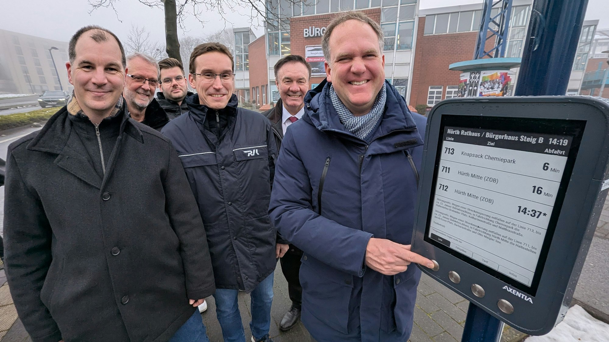 Das Foto zeigt eine Gruppe von Menschen vor dem digitalen Anzeigegerät an der Bushaltestelle.