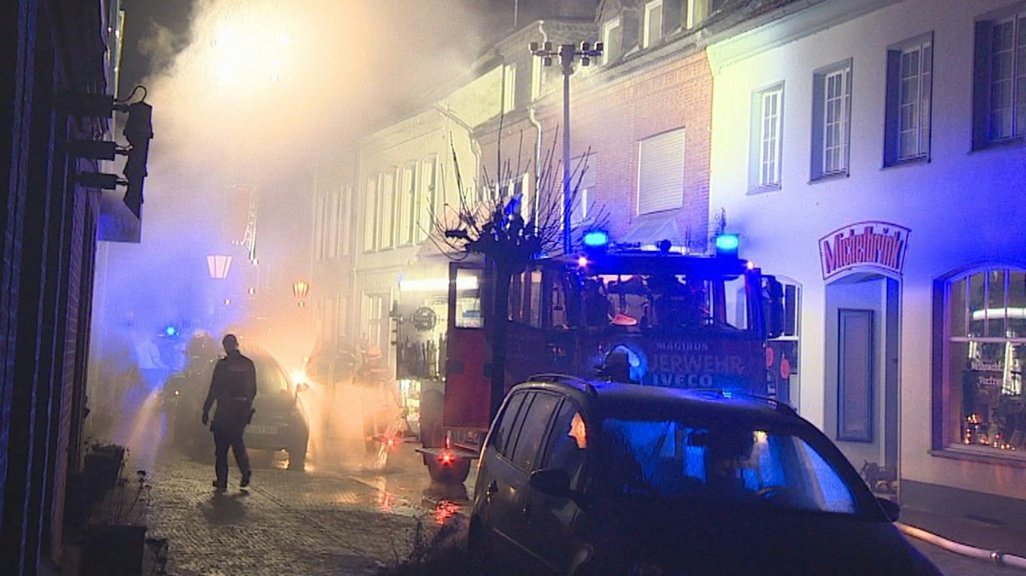 Einsatzkräfte bekämpfen einen Wohnungsbrand in Kalkar. Eine kleine Straße ist in der Nacht mit Rauch gefüllt, davor ein Feuerwehrauto mit Blaulicht.
