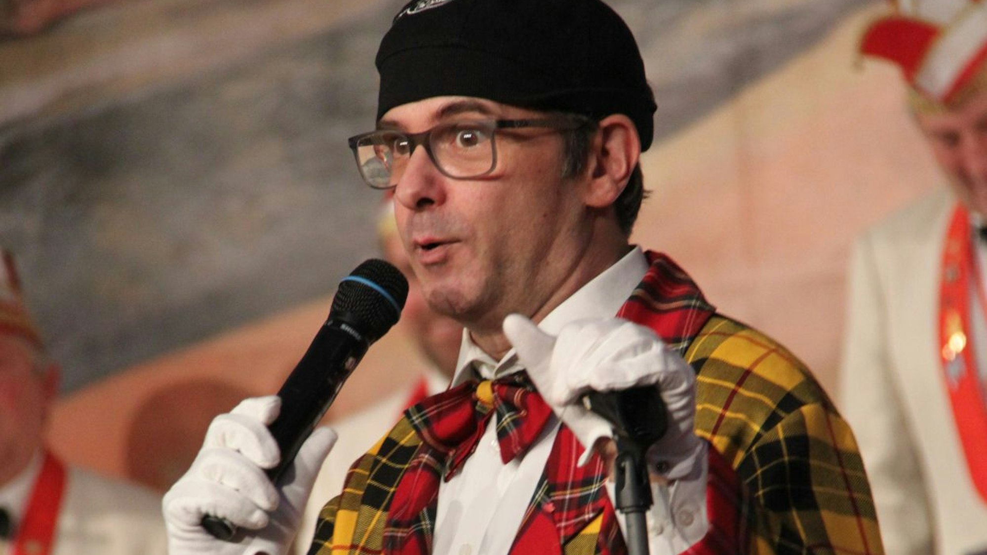 Marc Metzger in seiner Rolle als Blötschkopp: Er trägt ein schwarzes Hütchen, karierten Frack und weiße Handschuhe und spricht in ein Mikrofon.