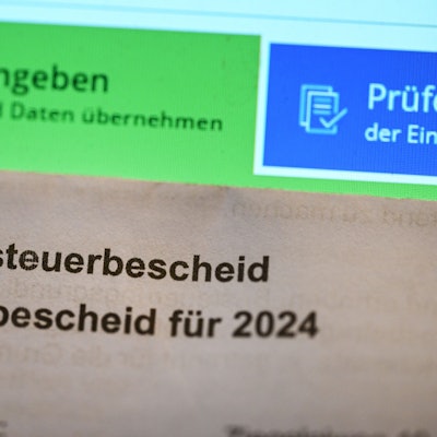 Ein Grundsteuerbescheid für 2024 wird vor einen Computerbildschirm gehalten, auf dem ein Formular der Steuerplattform Elster angezeigt wird. Die Zahl der Einsprüche gegen Steuerbescheide nimmt in Nordrhein-Westfalen deutlich zu.