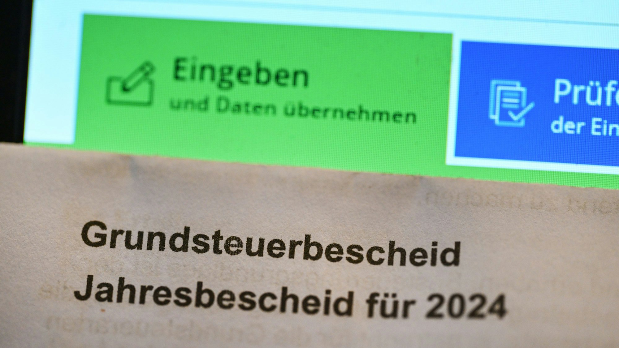 Ein Grundsteuerbescheid für 2024 wird vor einen Computerbildschirm gehalten, auf dem ein Formular der Steuerplattform Elster angezeigt wird. Die Zahl der Einsprüche gegen Steuerbescheide nimmt in Nordrhein-Westfalen deutlich zu.