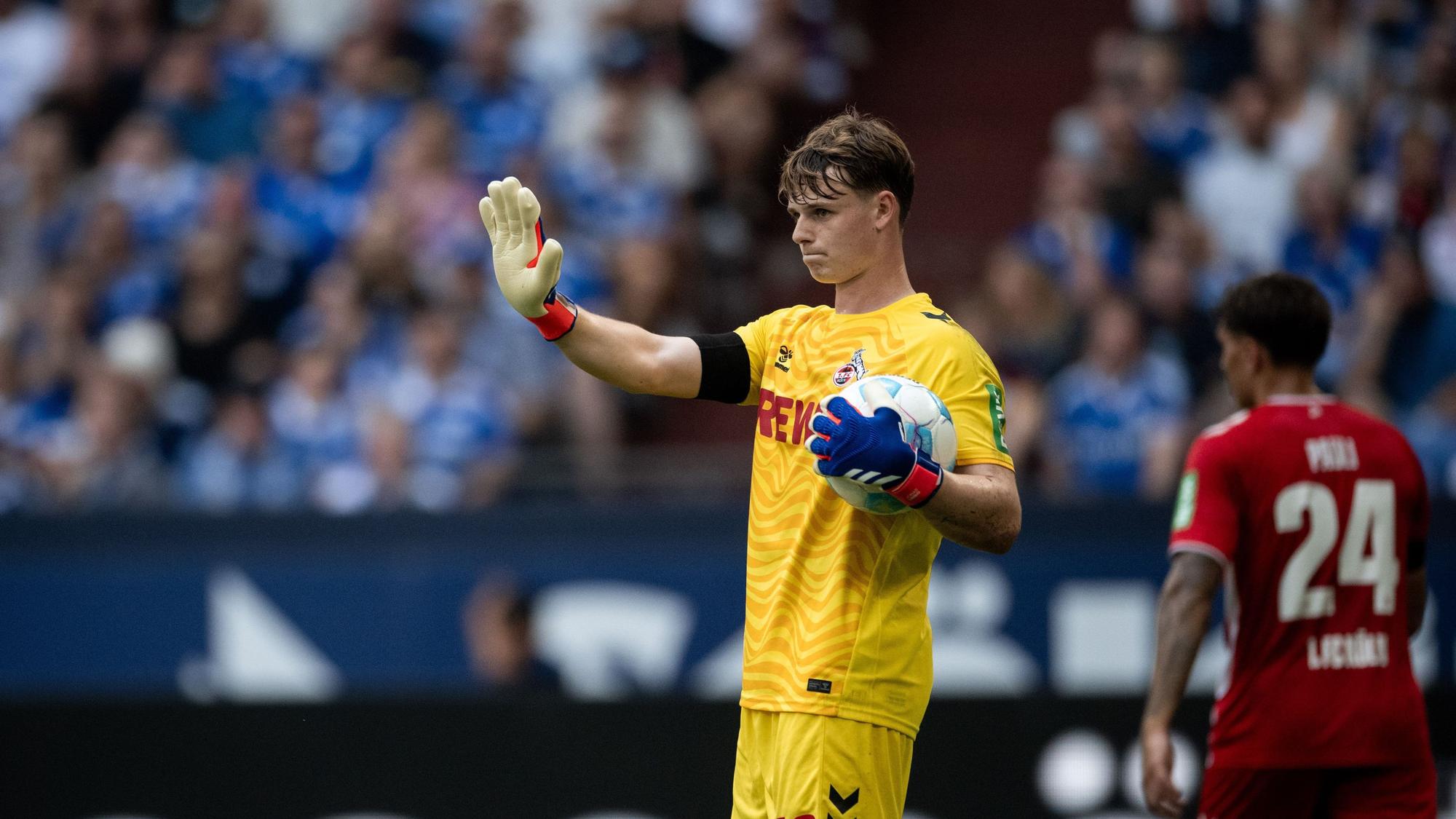 Jonas Urbig hält einen Ball in seinem Arm und die andere Hand hat er vor sich ausgestreckt.