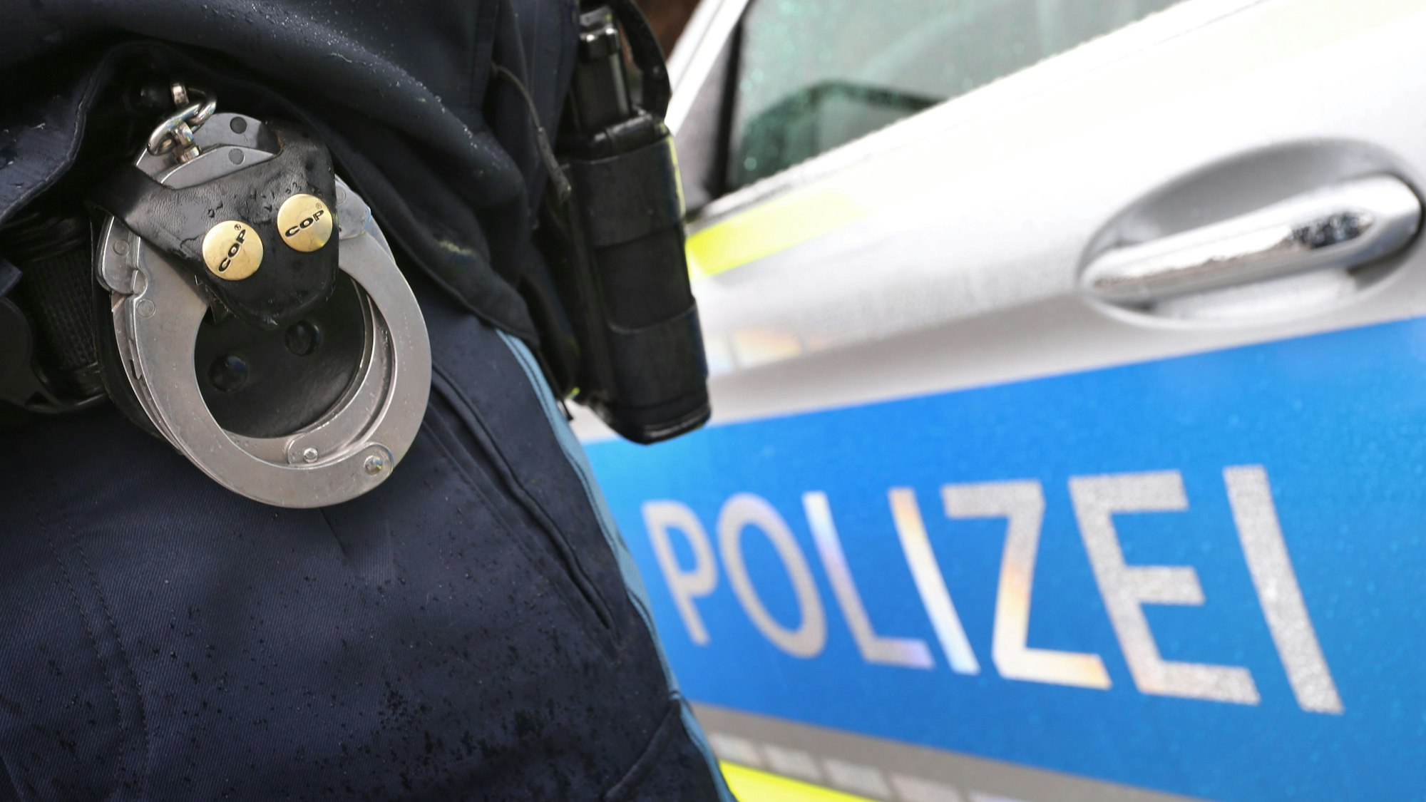 ARCHIV - 15.03.2019, Bayern, Kempten: Ein Polizist mit Handschellen und einer Pistole am Gürtel steht vor einem Streifenwagen.