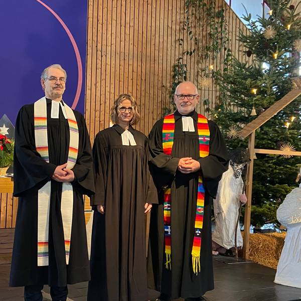 Zwei Männer und eine Frau stehen im schwarzen Ornat im Altarraum einer Kirche.
